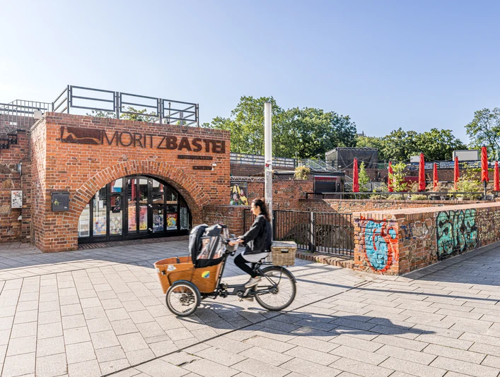Moritzbastei - Veranstaltungen in Leipzig Bild vom oberen Eingang zur Moritzbastei am Kurt-Masur-Platz in Leipzig, in der es ein Café und Restaurant gibt und in dem viele Veranstaltungen stattfinden, eine Fahrradfahrerin fährt durch das Bild