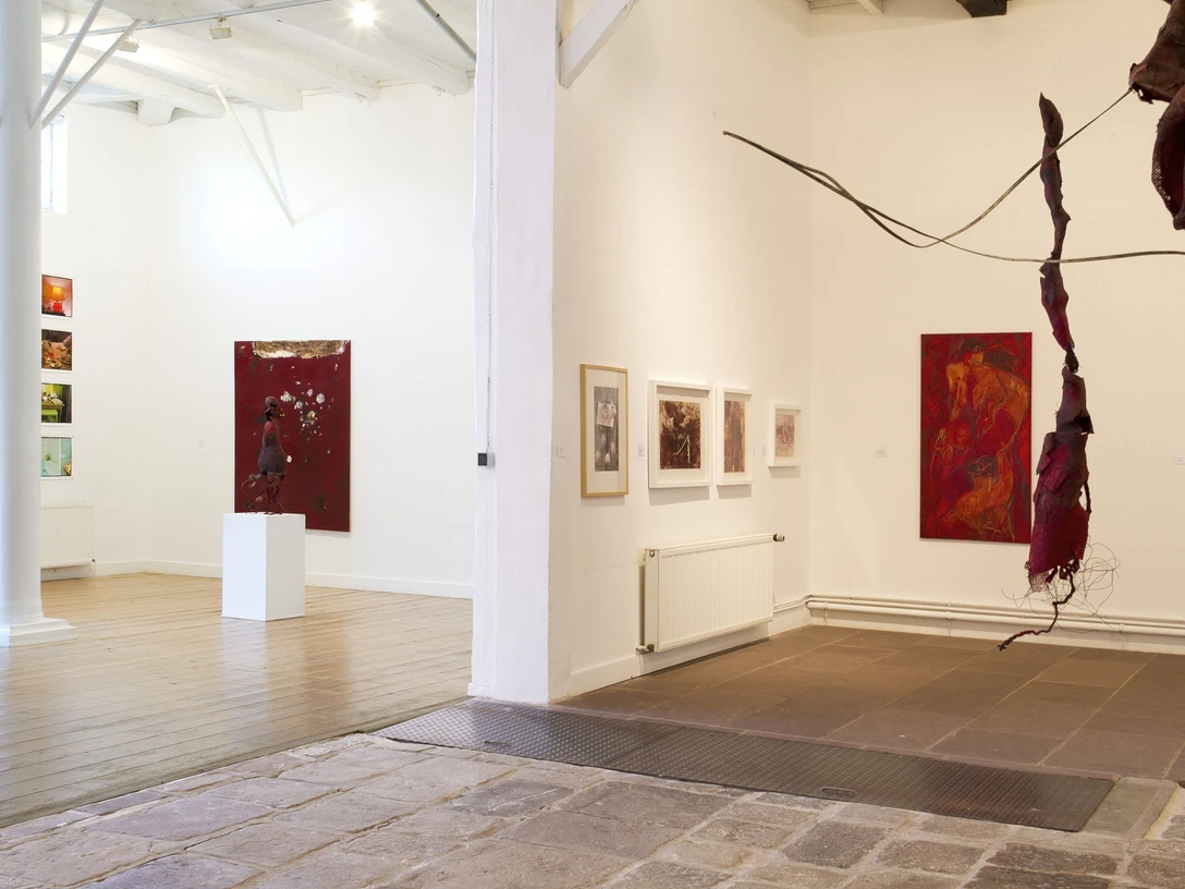 Kunstgalerie mit Holzboden und Gemälden an weißen Wänden, Struktur aus roten Stoffbahnen im Vordergrund.