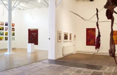 Koepkehaus_03.jpg Kunstgalerie mit Holzboden und Gemälden an weißen Wänden, Struktur aus roten Stoffbahnen im Vordergrund.