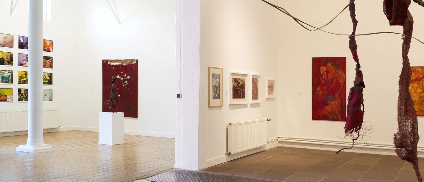 Koepkehaus_03.jpg Kunstgalerie mit Holzboden und Gemälden an weißen Wänden, Struktur aus roten Stoffbahnen im Vordergrund.