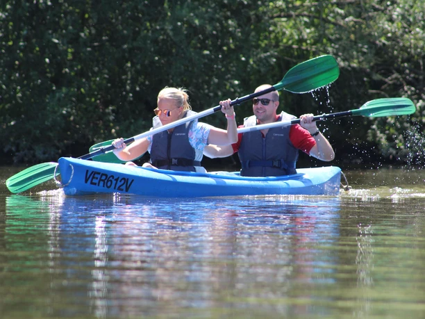Ein Mann und eine Frau paddeln in einem blauen Kanu durch ein ruhiges Gewässer, umgeben von grüner Natur.