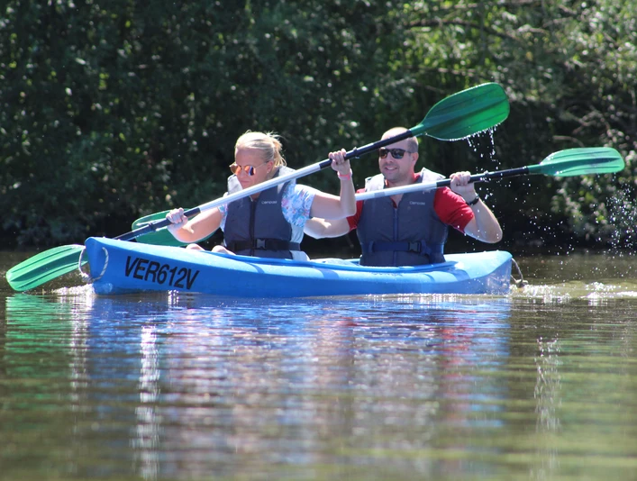 Kanutour Ein Mann und eine Frau paddeln in einem blauen Kanu durch ein ruhiges Gewässer, umgeben von grüner Natur.