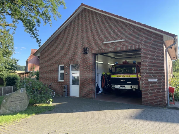 Freiwillige Feuerwehr Raven/Rolfsen Backsteingebäude mit offenem Feuerwehrtor, darin rotes Einsatzfahrzeug und Helme der Feuerwehr.