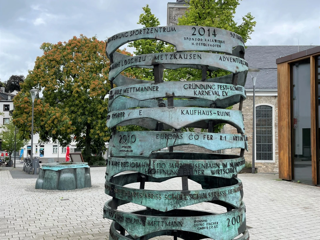 Erinneringe in Mettmann Eine skulpturale Bronze-Säule mit gravierten Jahreszahlen steht vor einer Kirche bei bewölktem Himmel.