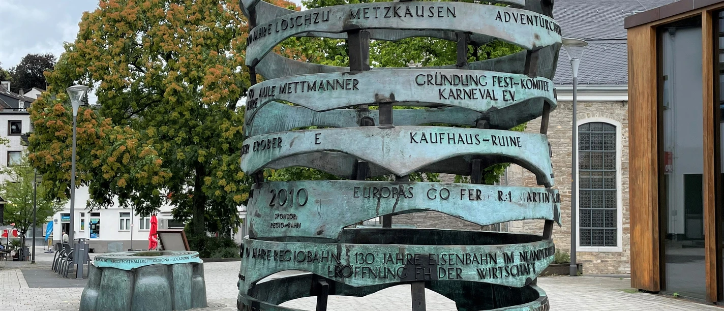 Erinneringe in Mettmann Eine skulpturale Bronze-Säule mit gravierten Jahreszahlen steht vor einer Kirche bei bewölktem Himmel.