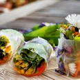 Kräuter-Tapas Das Bild zeigt drei frische Frühlingsrollen mit buntem Gemüse und essbaren Blüten auf einem Holztisch.