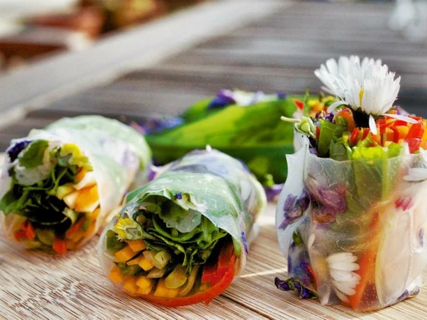 Kräuter-Tapas Das Bild zeigt drei frische Frühlingsrollen mit buntem Gemüse und essbaren Blüten auf einem Holztisch.