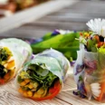 Kräuter-Tapas Das Bild zeigt drei frische Frühlingsrollen mit buntem Gemüse und essbaren Blüten auf einem Holztisch.
