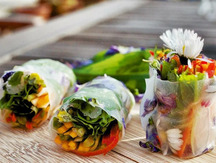 Kräuter-Tapas Das Bild zeigt drei frische Frühlingsrollen mit buntem Gemüse und essbaren Blüten auf einem Holztisch.