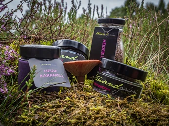 Verschiedene Salzsorten Das Bild zeigt eine natürliche Szene, in der verschiedene Gläser mit Heidelbeer- und Heidelblütenprodukten inmitten von Heidekraut arrangiert sind. Ein roter Pilz ergänzt das stimmungsvolle Naturbild.