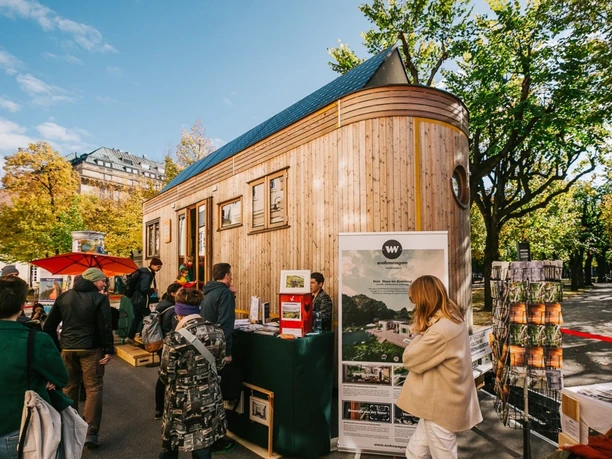 Green World Tour Cologne Several people surround a modern wooden tiny house that stands in a sunny spot.