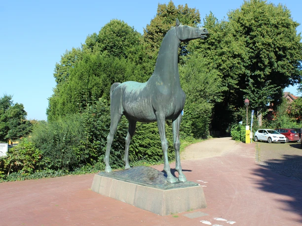 Bronzestatue eines Pferdes auf einem Podest, umgeben von üppigem Grün und gepflasterten Wegen.