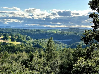 Zicht op een groen, heuvelachtig landschap met bossen en velden onder een gedeeltelijk bewolkte hemel; op de achtergrond is een stuwdam zichtbaar.