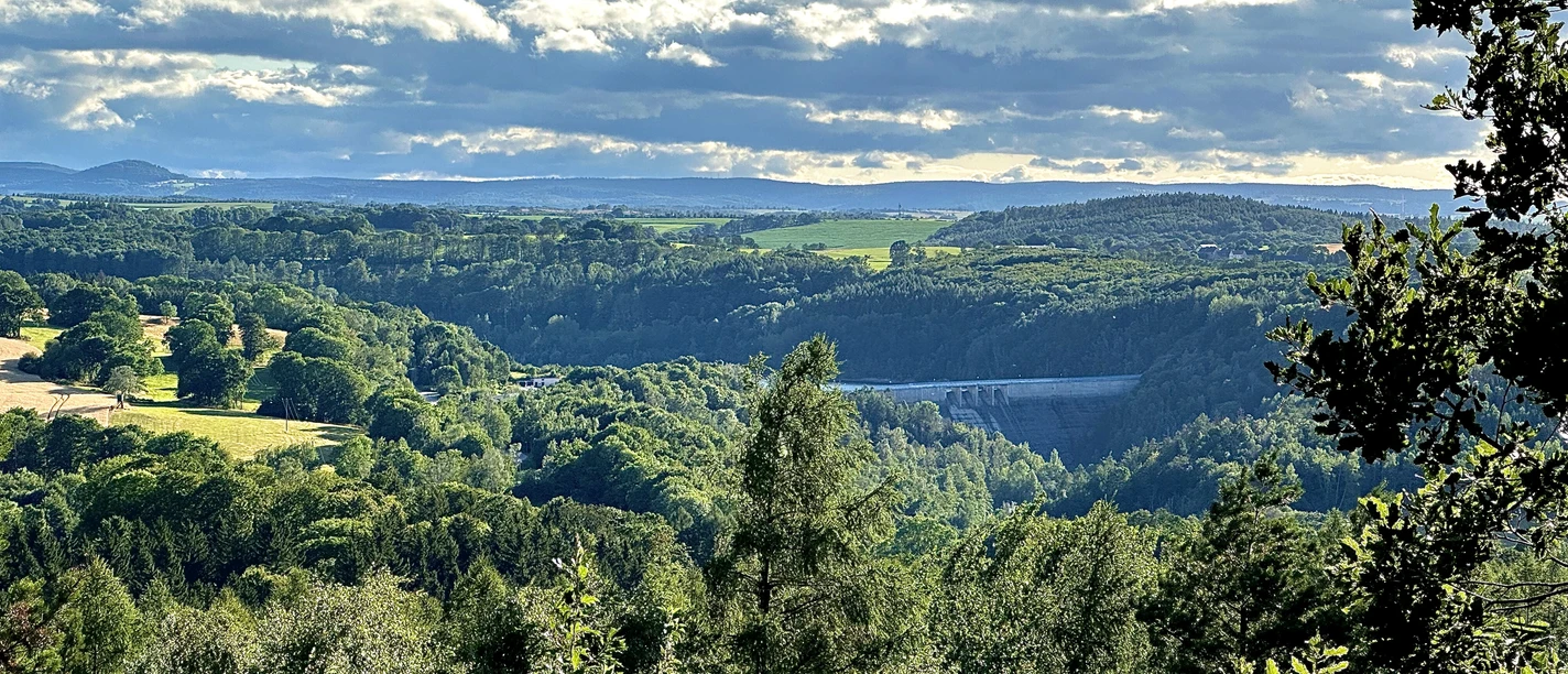 View of a green, hilly landscape with forests and fields under a partly cloudy sky; a dam is visible in the background.