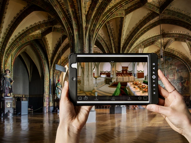 Handen die een tablet vasthouden met een virtuele voorstelling van een historische binnenhal.