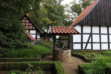 Heimatmuseum Versmold Fachwerkhäuser mit roten Ziegeldächern umgeben einen steinernen Brunnen in einem grünen Garten.
