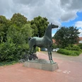 Wiehernder Hengst Bronzestatue eines Pferdes auf einem gepflasterten Platz, umgeben von Bäumen und Fahrrädern.
