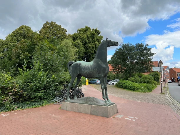 Wiehernder Hengst Bronzestatue eines Pferdes auf einem gepflasterten Platz, umgeben von Bäumen und Fahrrädern.