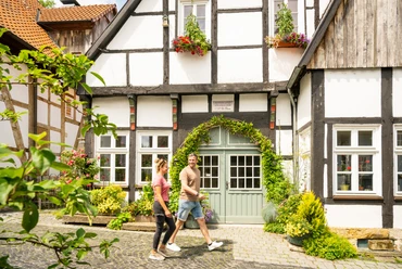 Tecklenburg-Altstadt-Tecklenburger Land, D. Ketz-2023-120.jpg Fachwerkhäuser mit Blumen und Grünpflanzen dekoriert, ein Paar spaziert eine gepflasterte Gasse entlang.