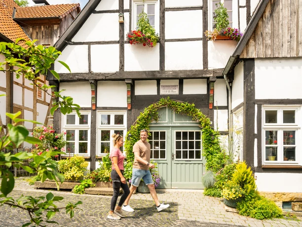 Tecklenburg-Altstadt-Tecklenburger Land, D. Ketz-2023-120.jpg Fachwerkhäuser mit Blumen und Grünpflanzen dekoriert, ein Paar spaziert eine gepflasterte Gasse entlang.