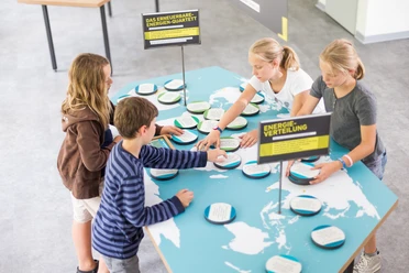 Stromwerkstatt Vier Kinder stehen um einen Tisch mit Weltkarten-Motiven und interagieren mit einem Energie-Quartett-Spiel.