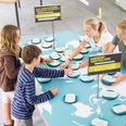 Stromwerkstatt Vier Kinder stehen um einen Tisch mit Weltkarten-Motiven und interagieren mit einem Energie-Quartett-Spiel.