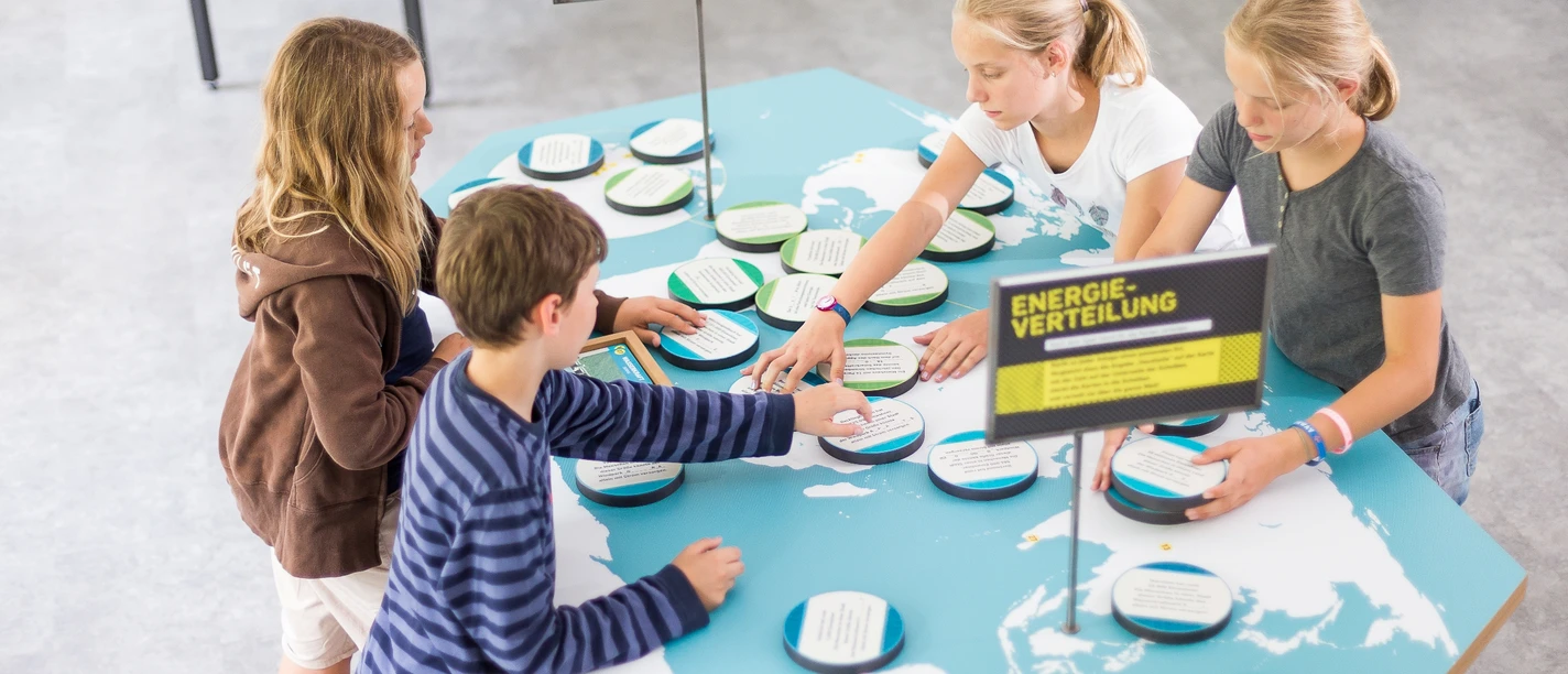 Stromwerkstatt Vier Kinder stehen um einen Tisch mit Weltkarten-Motiven und interagieren mit einem Energie-Quartett-Spiel.