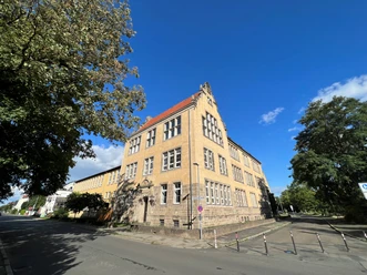 Dreistöckiges historisches Schulgebäude mit gelber Fassade und markantem Giebel unter blauem Himmel.