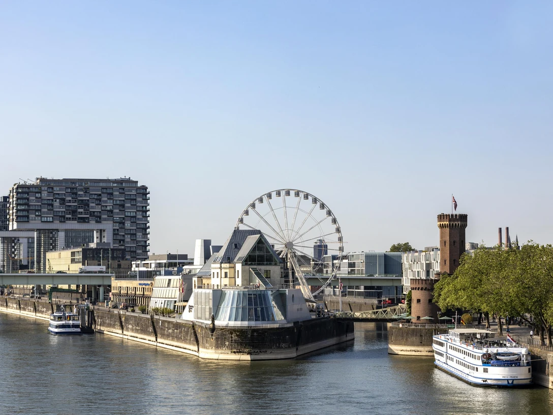 Cologne Ferris wheel Rheinufers in Köln mit dem Schokoladenmuseum im Vordergrund. Ein großes Riesenrad steht neben dem Museum, umgeben von modernen Gebäuden und einem historischen Turm. Das ruhige Wasser des Rheins und die vorbeifahrenden Schiffe verleihen der Szene eine malerische Atmosphäre.Bank of the Rhine in Cologne with the Chocolate Museum in the foreground. A large Ferris wheel stands next to the museum, surrounded by modern buildings and a historic tower. The calm waters of the Rhine and the passing ships lend the scene a picturesque atmosphere.