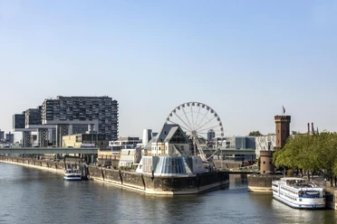 Kölner Riesenrad Rheinufers in Köln mit dem Schokoladenmuseum im Vordergrund. Ein großes Riesenrad steht neben dem Museum, umgeben von modernen Gebäuden und einem historischen Turm. Das ruhige Wasser des Rheins und die vorbeifahrenden Schiffe verleihen der Szene eine malerische Atmosphäre.