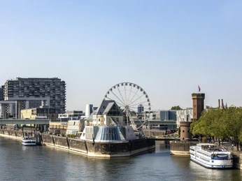 Kölner Riesenrad Rheinufers in Köln mit dem Schokoladenmuseum im Vordergrund. Ein großes Riesenrad steht neben dem Museum, umgeben von modernen Gebäuden und einem historischen Turm. Das ruhige Wasser des Rheins und die vorbeifahrenden Schiffe verleihen der Szene eine malerische Atmosphäre.Bank of the Rhine in Cologne with the Chocolate Museum in the foreground. A large Ferris wheel stands next to the museum, surrounded by modern buildings and a historic tower. The calm waters of the Rhine and the passing ships lend the scene a picturesque atmosphere.