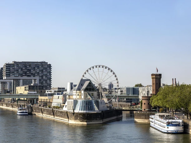 Kölner Riesenrad Rheinufers in Köln mit dem Schokoladenmuseum im Vordergrund. Ein großes Riesenrad steht neben dem Museum, umgeben von modernen Gebäuden und einem historischen Turm. Das ruhige Wasser des Rheins und die vorbeifahrenden Schiffe verleihen der Szene eine malerische Atmosphäre.