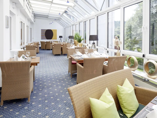 Waldhotel Baerenstein GmbH_Wintergarten.jpg Heller Wintergarten eines Waldhotels mit Rattanmöbeln; Blick auf grüne Bäume durch große Fenster.