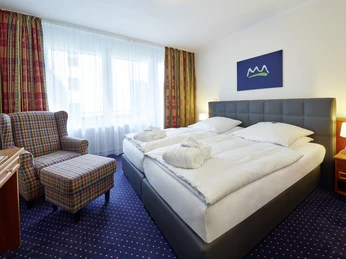 Waldhotel Bärenstein Doppelzimmer.jpg Helles Doppelzimmer mit zwei Betten, gepunktetem Teppich, Sessel, Tisch und naturinspiriertem Kunstwerk.