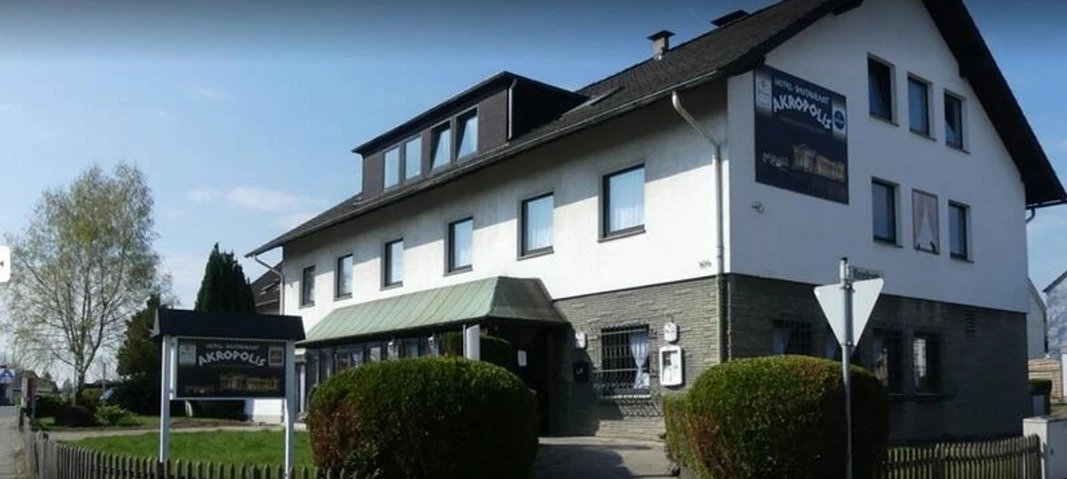 Zweistöckiges Gebäude mit Akropolis Restaurant-Schild, weißer Fassade, grünem Vorbau und Holzzaun.