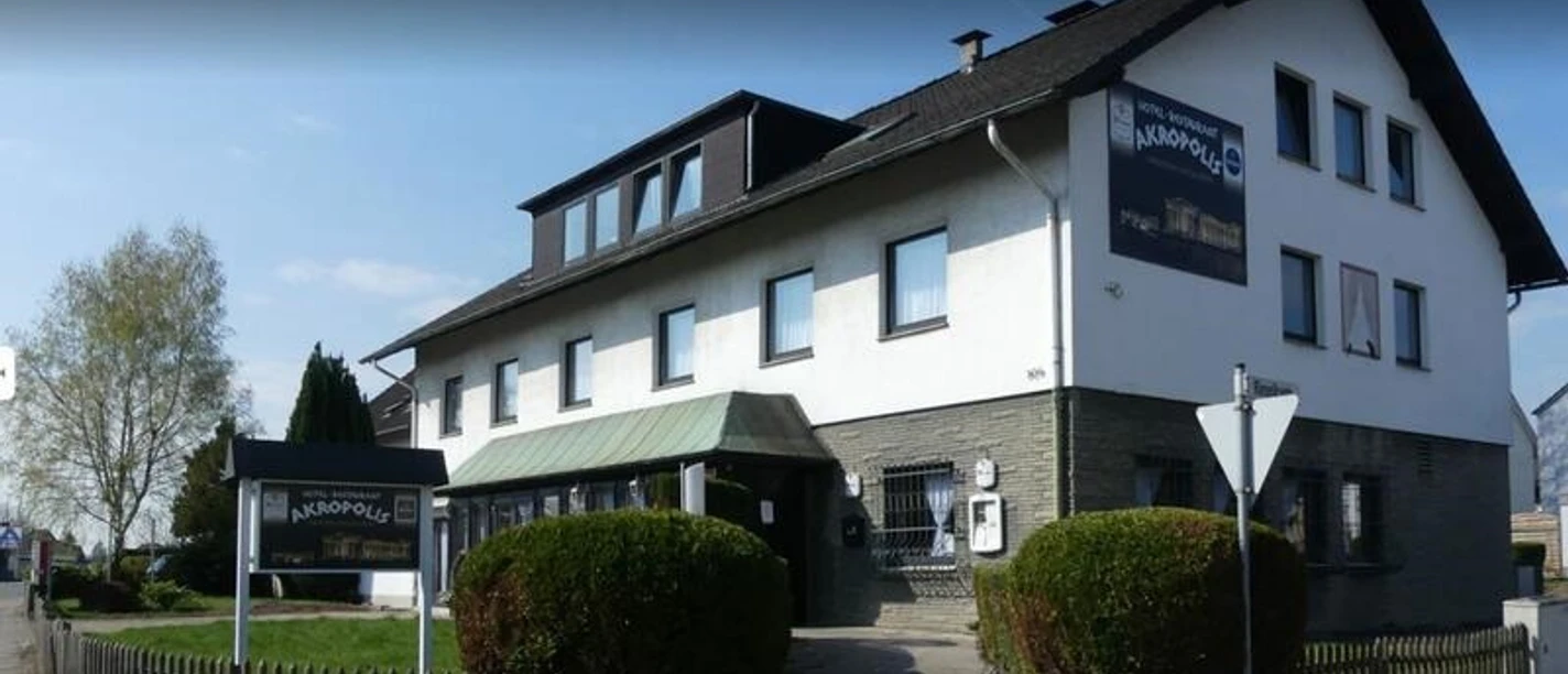 Zweistöckiges Gebäude mit Akropolis Restaurant-Schild, weißer Fassade, grünem Vorbau und Holzzaun.