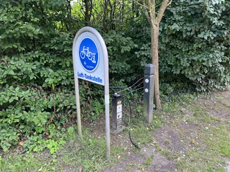 Fahrrad-Lufttankstelle am Waldrand mit Manometer und Reifenventil, umgeben von grünem Laub.