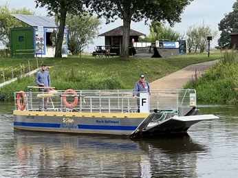 Solar Fähre Otersen-Westen Eine Solarfähre überquert ruhig einen Fluss, wobei zwei Personen an Deck stehen und das Ufer erkennbar ist.