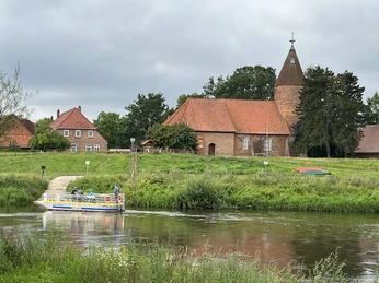 Kirche Westen Backsteinkirche mit Spitzdach und Turm an Flussufer, kleines Boot am Steg, umgeben von Grünflächen.
