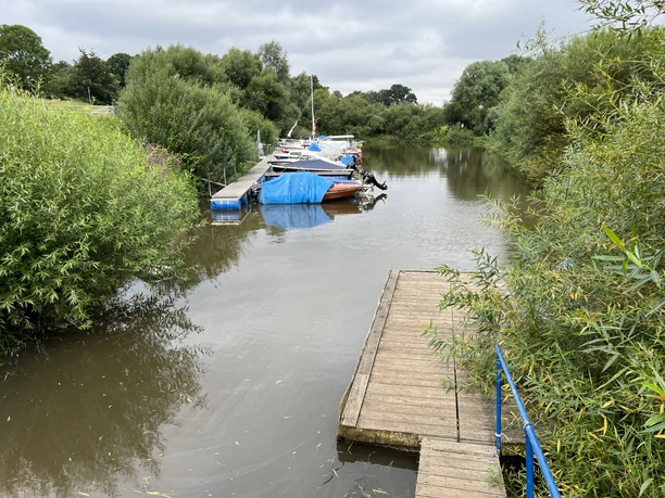 Mehrere kleine Boote entlang eines Holzstegs in einem ruhigen, von Bäumen gesäumten Fluss bei bewölktem Himmel.