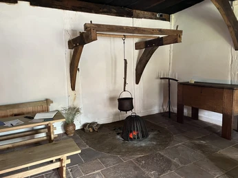 Ein rustikaler Raum mit einem eisernen Kochtopf über einer offenen Feuerstelle, umgeben von Holzmöbeln.