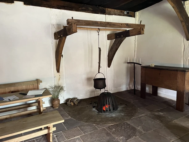 Ein rustikaler Raum mit einem eisernen Kochtopf über einer offenen Feuerstelle, umgeben von Holzmöbeln.