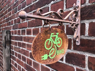 Ein rostfarbenes Schild mit grünem E-Bike-Symbol hängt an einer roten Backsteinwand.