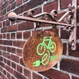 E-Bike Ladestation Ein rostfarbenes Schild mit grünem E-Bike-Symbol hängt an einer roten Backsteinwand.