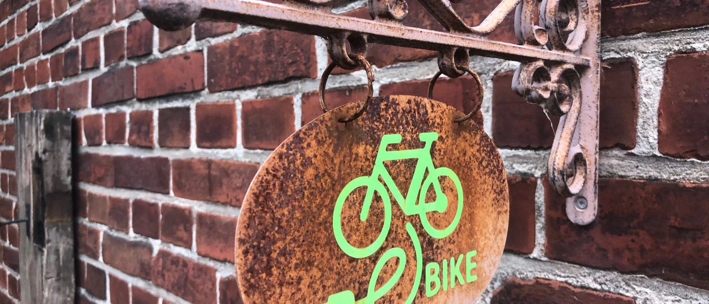 E-Bike Ladestation Ein rostfarbenes Schild mit grünem E-Bike-Symbol hängt an einer roten Backsteinwand.