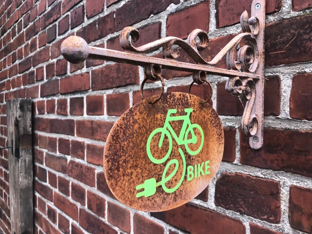 E-Bike Ladestation Ein rostfarbenes Schild mit grünem E-Bike-Symbol hängt an einer roten Backsteinwand.