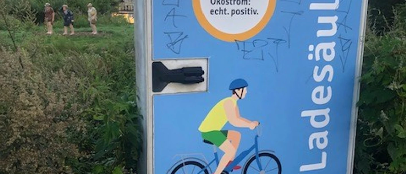 Eine Elektroladesäule mit Graffiti, daneben üppige Vegetation und Hintergrundpersonen.