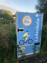 Mühlenwall.jpg Eine Elektroladesäule mit Graffiti, daneben üppige Vegetation und Hintergrundpersonen.