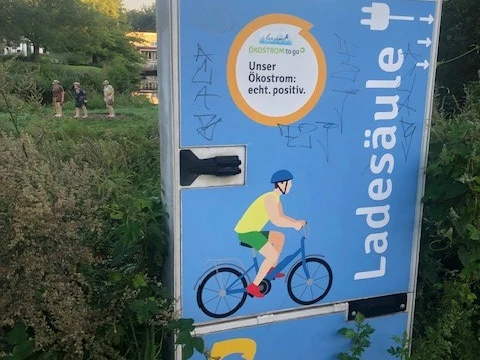 Mühlenwall.jpg Eine Elektroladesäule mit Graffiti, daneben üppige Vegetation und Hintergrundpersonen.