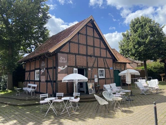 Flying Barista Ein rotes Fachwerkhaus mit Sitzgelegenheiten im Freien und Sonnenschirmen unter blauem Himmel.